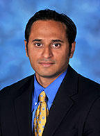 Man wearing a blue shirt, black suit and yellow tie posing for a portrait photo with blue background.