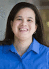 A woman wearing a blue-collared shirt smiling against a brown background. She has shoulder-length dark brown hair. 